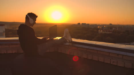 A-programmer-with-a-laptop-in-headphones-sits-on-the-roof-and-writes-code-at-sunset-and-listens-to-music.-Remote-work-freelancer.-Freedom-to-work.-Typing-on-a-keyboard-at-sunset-with-a-view-of-the-city.